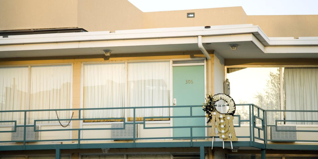 A simple motel balcony with room 306 marked, featuring a memorial wreath placed near the railing in front of a pale blue door.