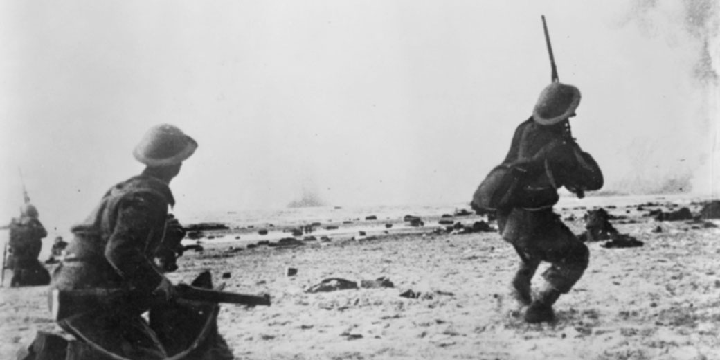 A black-and-white photo of soldiers advancing on a beach during battle. They wear helmets and carry rifles with bayonets attached.