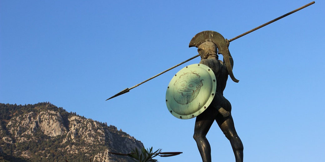 Statue of an ancient Greek hoplite holding a spear and shield with a Gorgon face, set against a clear sky and rocky hillside.