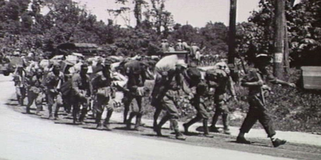 A line of weary soldiers or prisoners marches along a rural road, heavily burdened with gear, under armed guard during wartime.