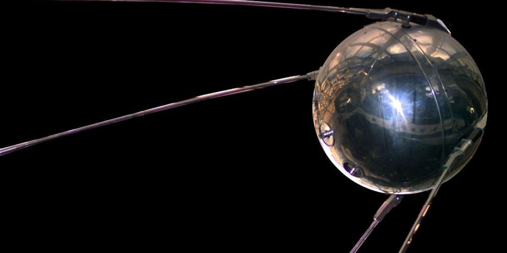 A shiny metallic sphere with long antennae extends outward against a black background, showing the historic Sputnik satellite.