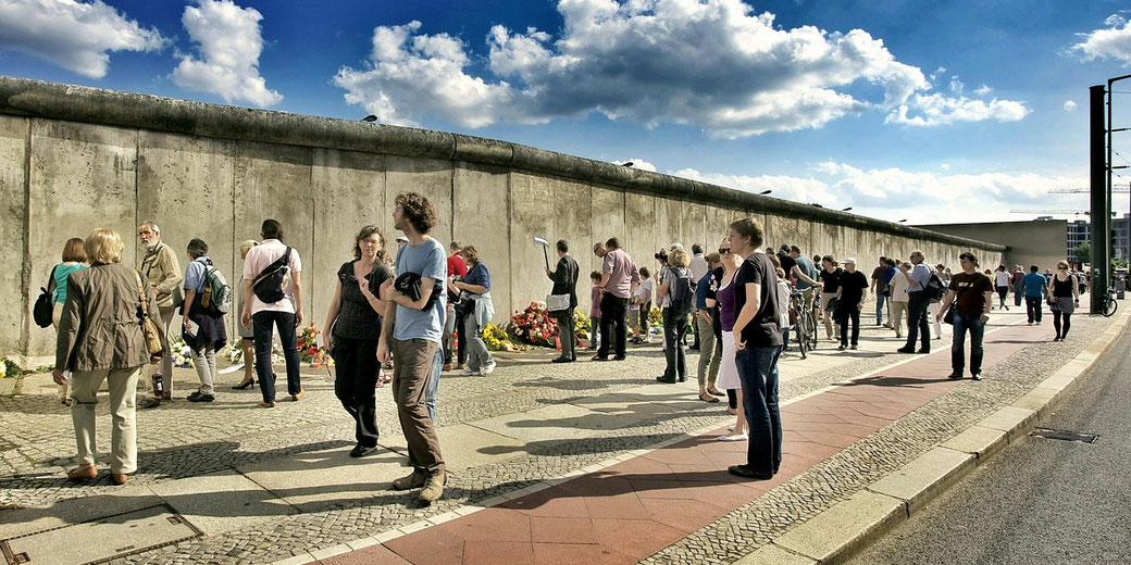 Crowd gathers beside a historic concrete wall, with some placing flowers in remembrance on a bright day under a partly cloudy sky.