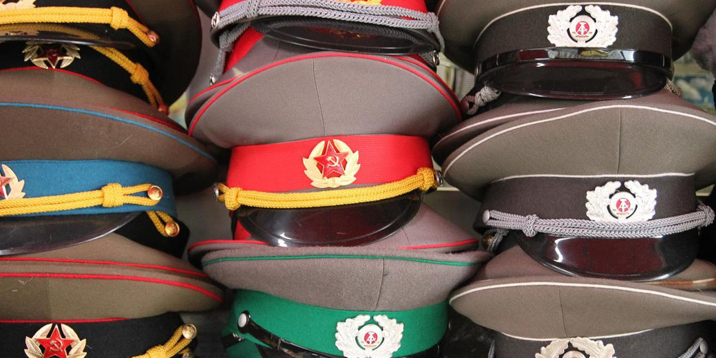 A stack of Soviet and East German military hats with colorful bands and insignia is tightly arranged on display.