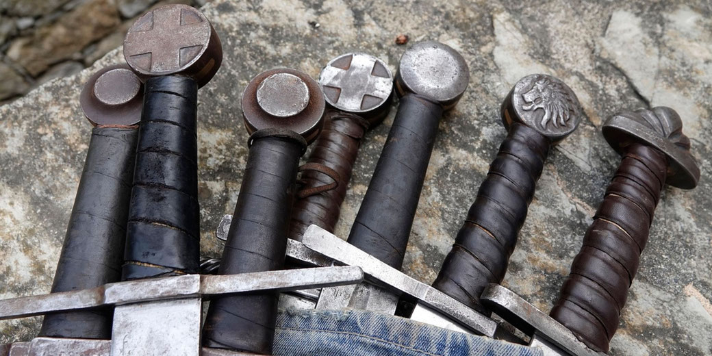 Several medieval-style swords with wrapped grips and distinct pommel designs, including crosses and a lion emblem, resting on a stone surface with part of a sheath visible.