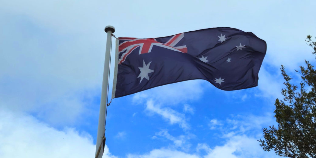 The Australian flag waves on a tall pole against a cloudy sky with patches of blue and nearby tree branches.
