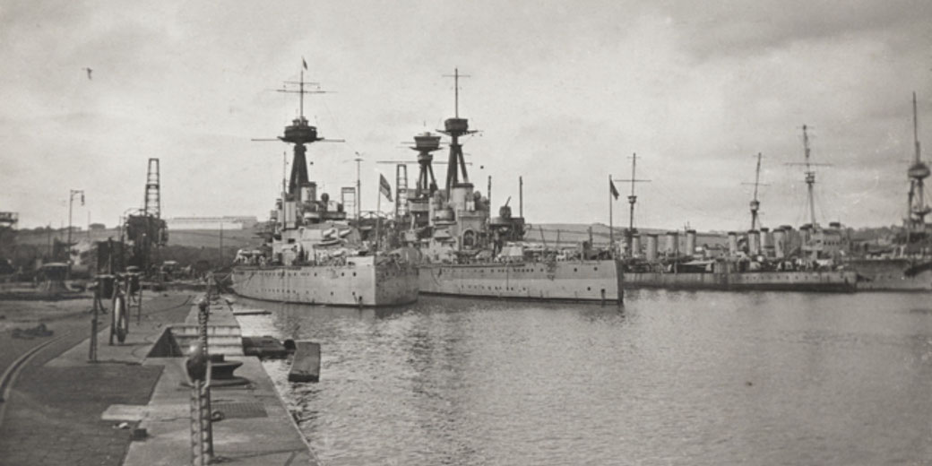 Several large warships are docked side by side in a harbor, with cranes and equipment visible along the pier.