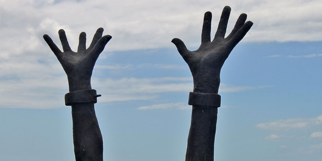 Dark sculpture of two raised hands with cuffs reaching upward against a cloudy sky.