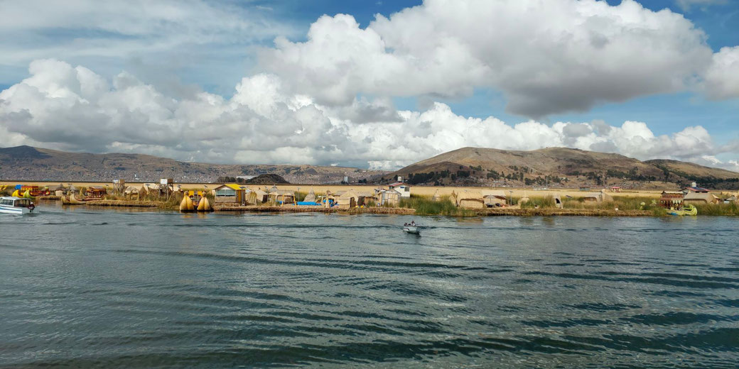 A floating village on Lake Titicaca, with reed houses and boats. The calm water reflects the sky, while distant hills and clouds add depth to the scene. A small boat is seen moving across the water.