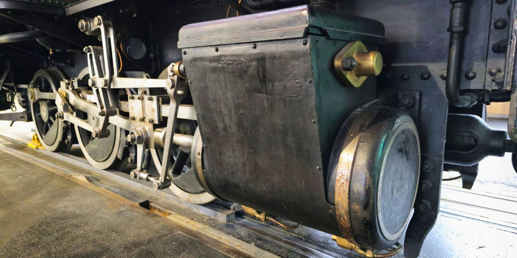 A close-up view of a steam locomotive's mechanical components, including large driving wheels, rods, and a worn metal casing with brass fittings.