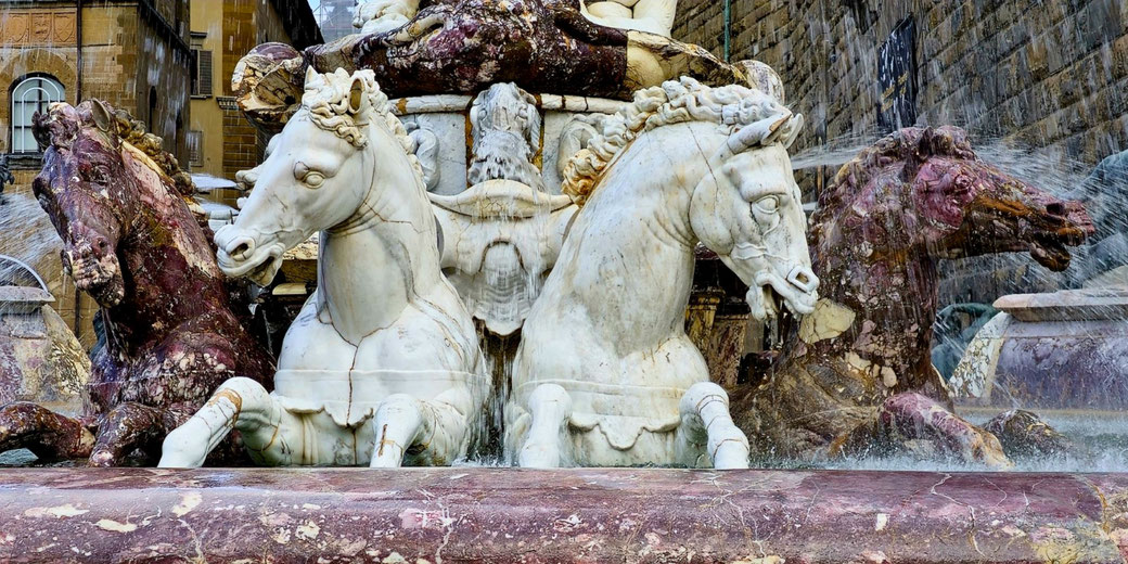 A marble fountain features four rearing horses, two white and two dark, with water spraying from their mouths and cascading down behind them.