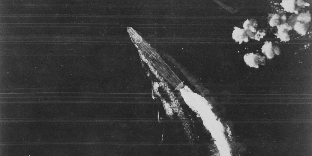 Black and white image of a missile or rocket rising diagonally with a bright exhaust plume, smoke trail, and scattered bursts nearby, captured from above with grainy texture.
