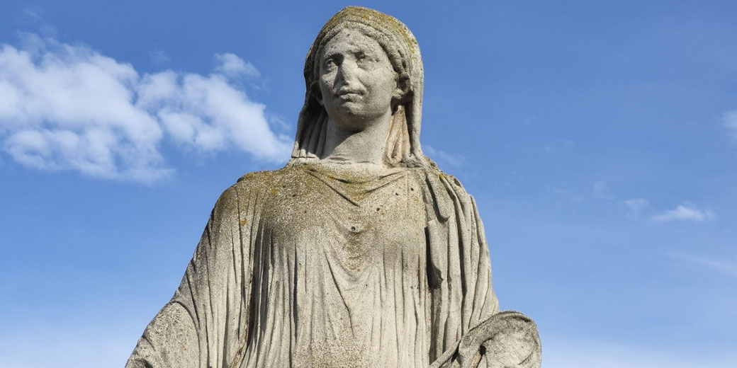 Weathered stone statue of a veiled woman in draped robes, holding a round object, set against a bright blue sky with scattered clouds.