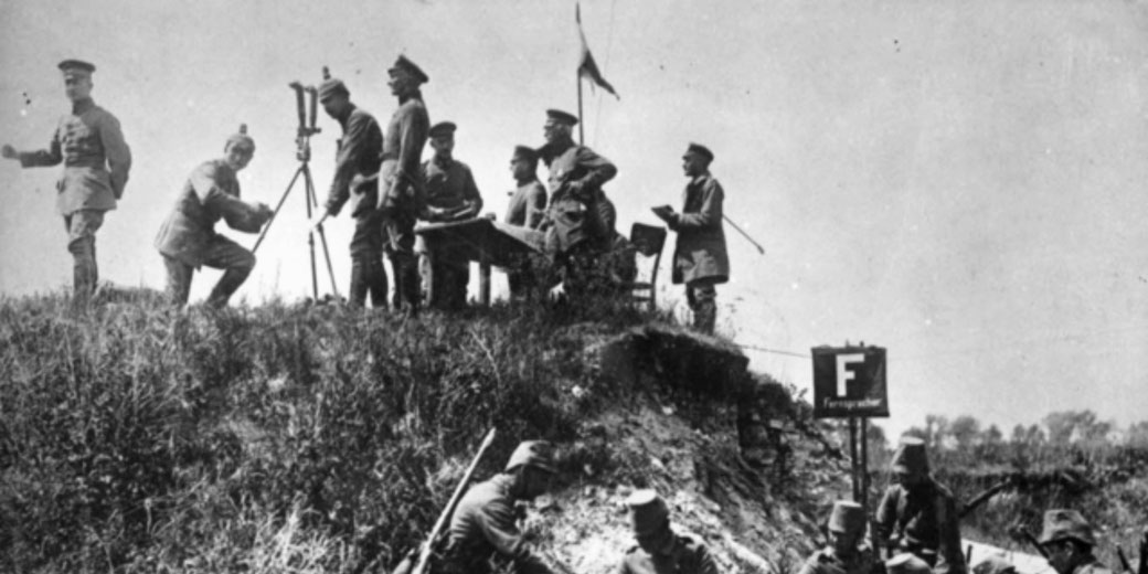 World War I soldiers set up equipment and gather around a table on a grassy hill, with some troops in a trench below near a sign labeled “F Fernsprecher.”