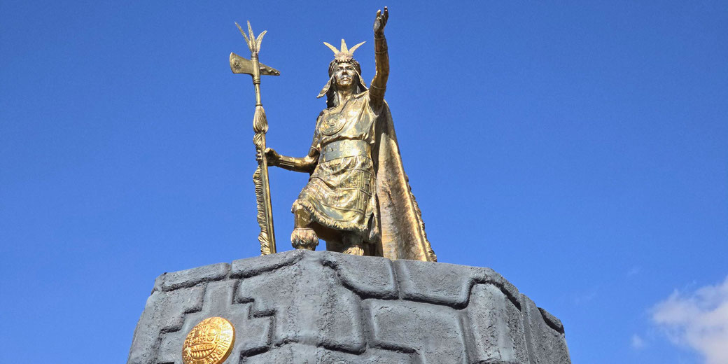 A golden statue of Pachacuti, the great Inca ruler, stands atop a stone pedestal in Cusco, Peru. Holding a ceremonial axe and wearing a regal cape, he raises his hand against a clear blue sky.