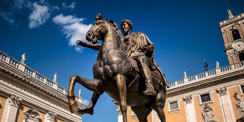 Bronze equestrian statue of a Roman figure in a plaza, framed by classical buildings under a bright blue sky.