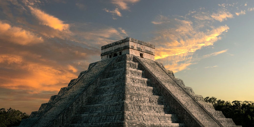 Mayan pyramid of Chichén Itzá in Mexico, bathed in the warm hues of a sunset. The stone structure stands tall, with its steep steps leading to a small temple at the top.