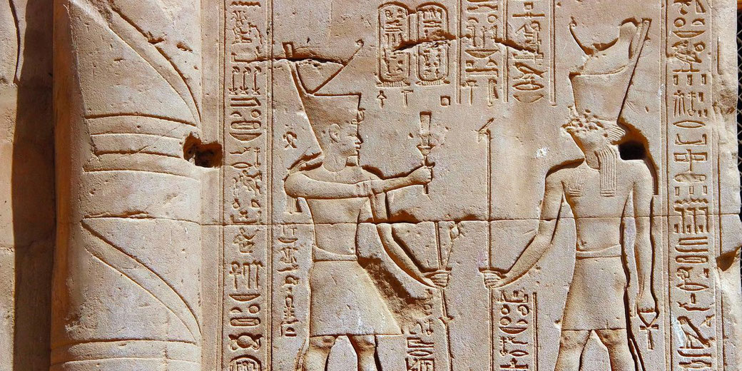 Ancient Egyptian wall carving shows two pharaohs with tall crowns holding staffs, surrounded by detailed hieroglyphs on stone.