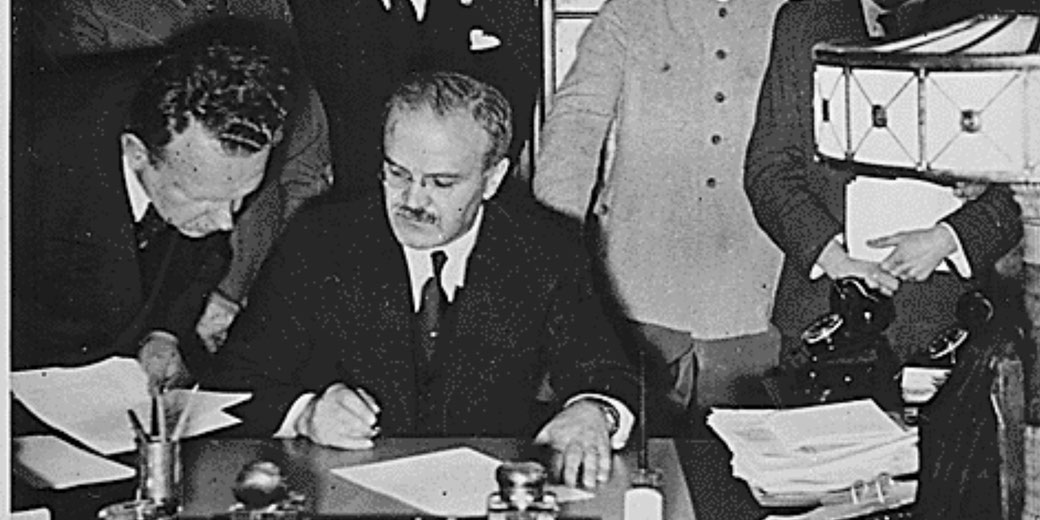 A man in a dark suit signs a document at a desk while several others look on, suggesting a formal or political agreement is being made.