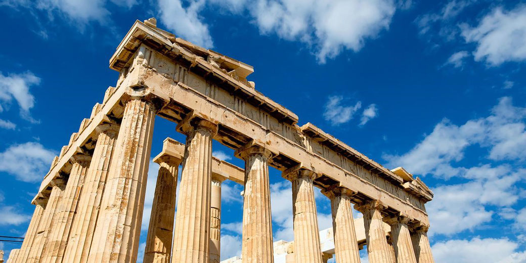 Ancient Greek temple ruins with tall stone columns under a bright blue sky with scattered clouds.
