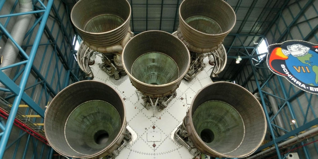 Low angle view of five large rocket engine nozzles on the base of a Saturn V stage inside a steel hangar, with blue support beams and overhead lights.