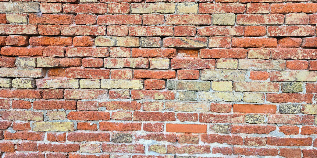 A close-up of an aged brick wall with red and beige bricks arranged in an irregular pattern. Some bricks appear worn or protruding, while the mortar shows signs of erosion, adding texture and character.