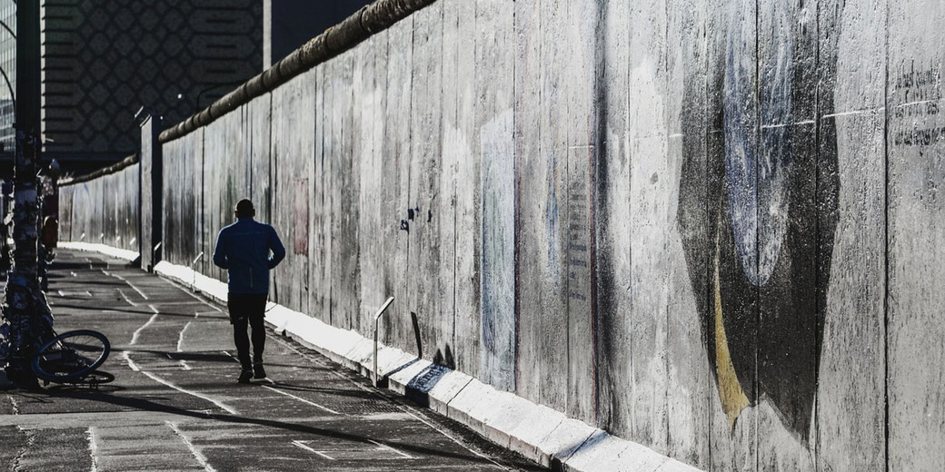 A lone runner moves alongside the graffiti-covered Berlin Wall in a shadowy urban setting.