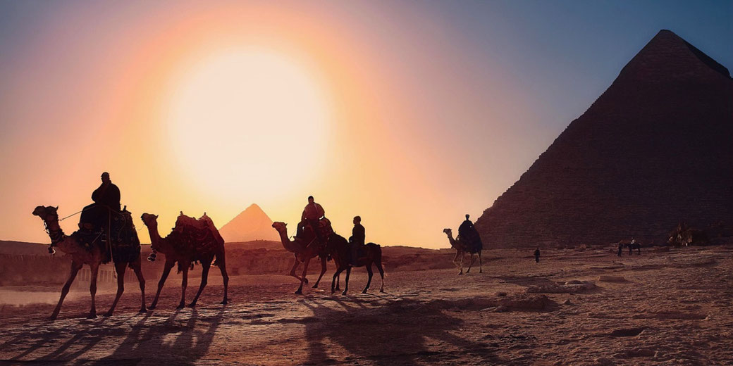 A stunning sunset over the Egyptian pyramids, with silhouetted camels and riders in the foreground. The warm glow highlights the desert landscape and the grandeur of the pyramids.
