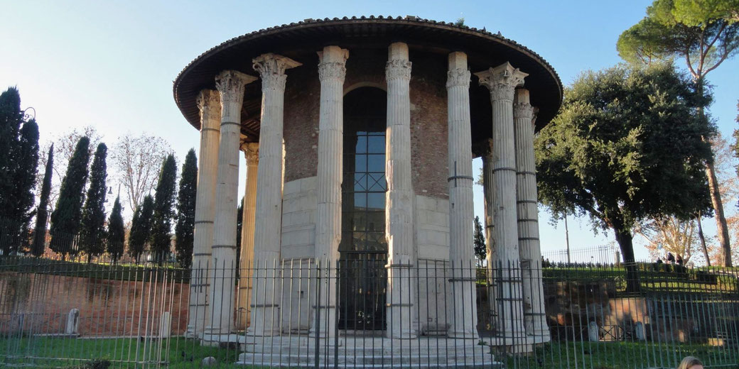 Circular Roman temple with Corinthian columns and a tiled roof, enclosed by a metal fence, surrounded by trees and greenery.