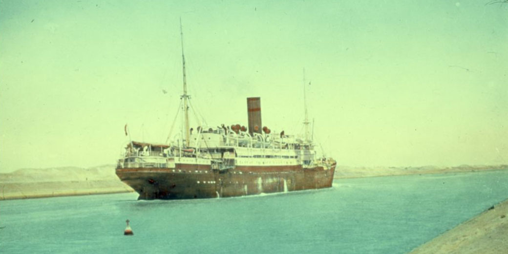 A steamship with a single funnel sails through a narrow canal flanked by sandy banks under a pale sky.