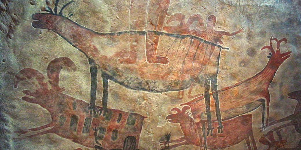 A prehistoric cave painting depicting animals and human figures in red and black pigments. The artwork portrays hunting scenes, likely created by early humans as part of ritual or storytelling.