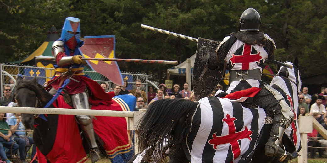 Two knights jousting at a medieval reenactment. One knight wears blue and red, while the other wears black and white with a red cross.