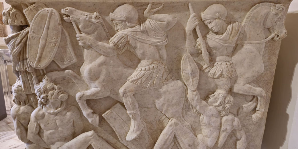 Marble relief showing Roman soldiers on horseback in battle, trampling and attacking fallen, defeated figures with shields and spears.
