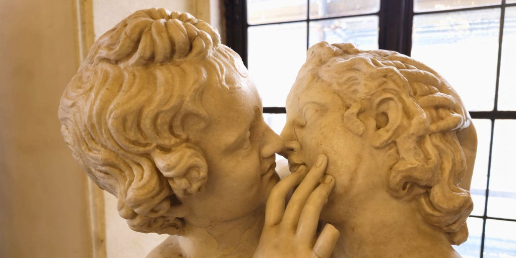 A close-up of a marble sculpture depicting two cherubs or young figures in an intimate moment, with one gently touching the other's lips as they lean in closely, their faces nearly touching.