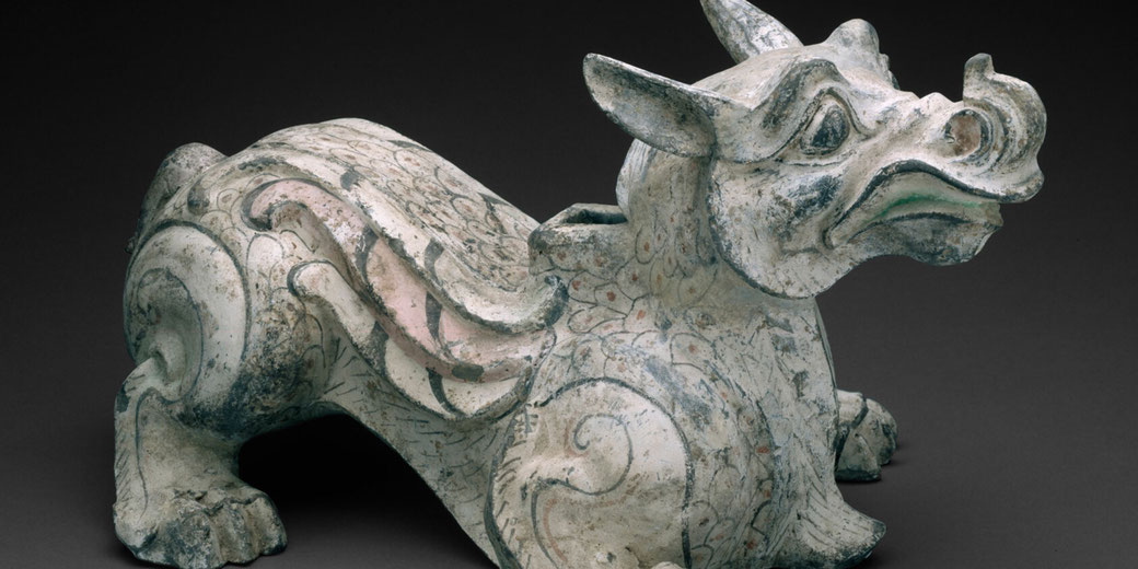 An ancient Chinese tomb figure of a winged beast, possibly from the Han Dynasty. It has a mythical appearance, with intricate swirling patterns and faded pigments on its surface.