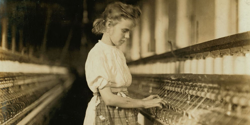 A young girl in a factory operates a spinning machine, likely from the early 20th century. She wears a worn dress and apron.