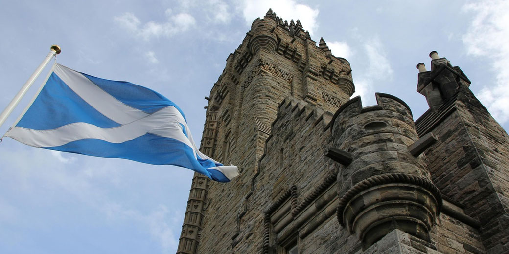 The Wallace Monument, a tall stone tower with intricate architecture, against a cloudy sky. A Scottish flag waves in the foreground.