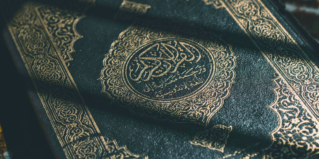 Close-up of the Quran cover with gold Arabic calligraphy and intricate patterns, partially shadowed and richly textured.