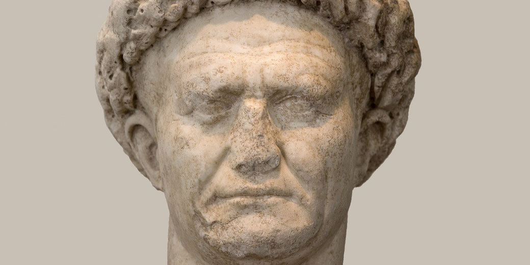 Weathered marble bust of a stern-faced man with curly hair, showing age and texture against a plain background.