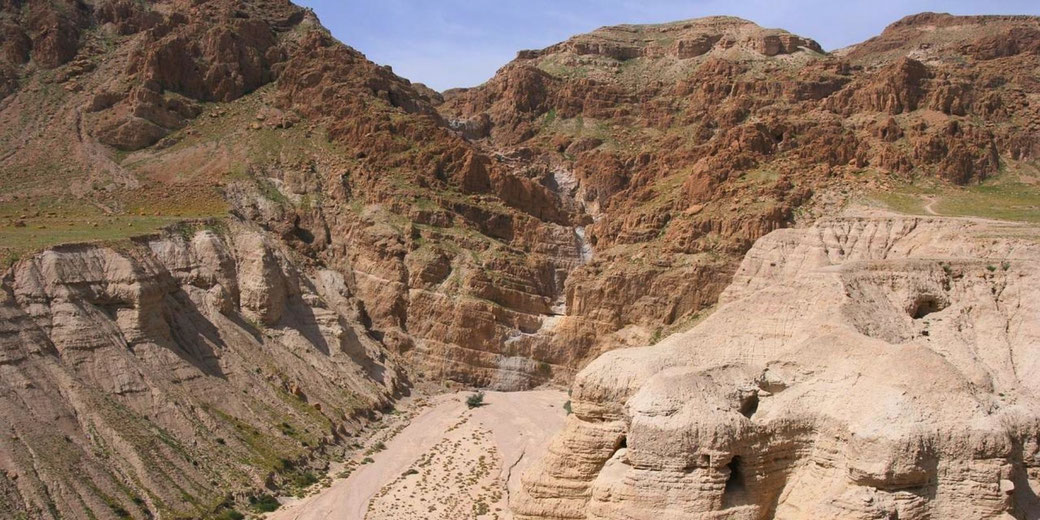 A rugged desert landscape with steep, eroded cliffs and a dry riverbed. The terrain is rocky with sparse vegetation. A cave is visible in the foreground, hinting at possible historical significance.