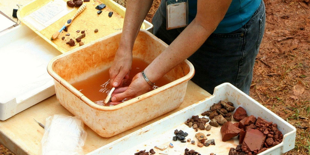 A person carefully cleans archaeological artifacts in a water tub, surrounded by trays of dirt-covered stones and tools.
