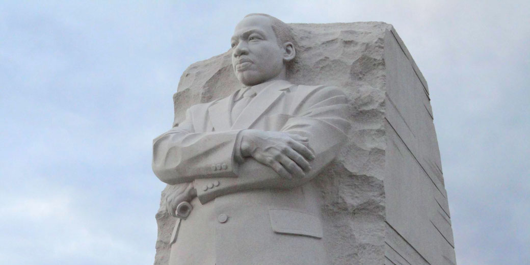 A large stone statue of a man in a suit with crossed arms is carved from a rock slab, looking forward with a serious expression.