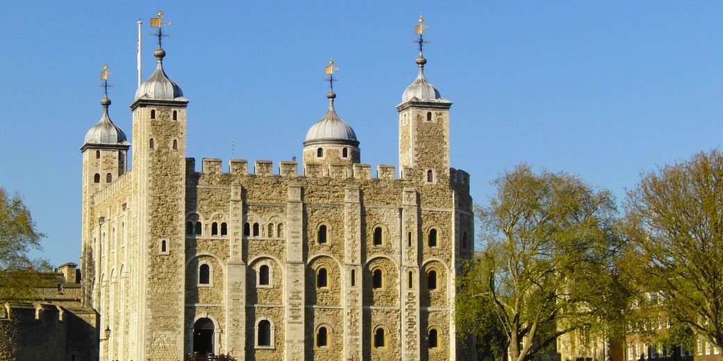 The Tower of London, a historic stone fortress in England. The medieval castle has four turrets, arched windows, and a crenelated parapet, set against a clear blue sky with trees nearby.