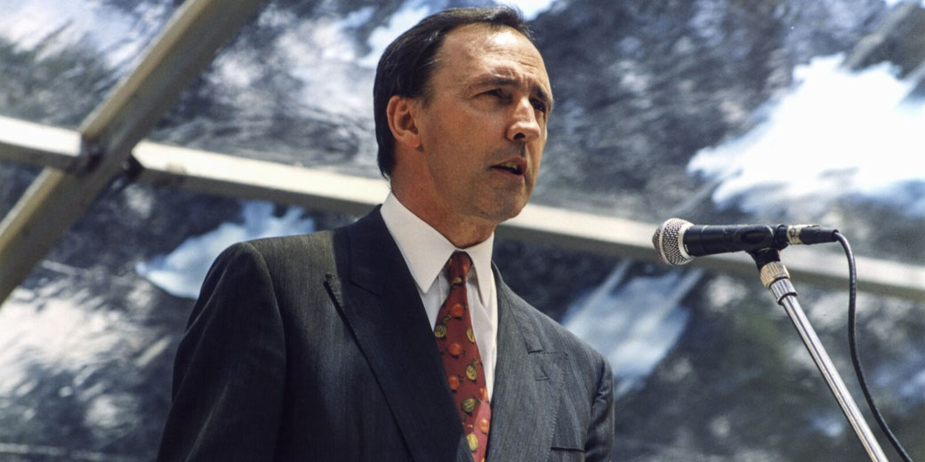A man in a dark suit and red tie addresses a crowd from behind a microphone under a glass canopy.