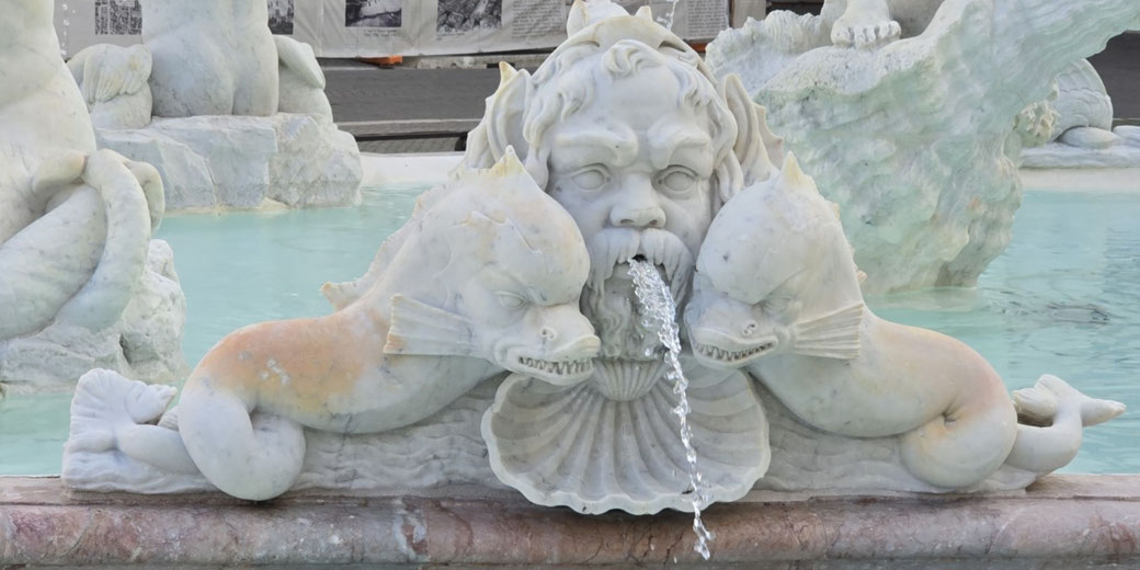 Ornate marble fountain featuring a bearded face and two sea creatures, all spouting water into a pool, with intricate shell and aquatic details.