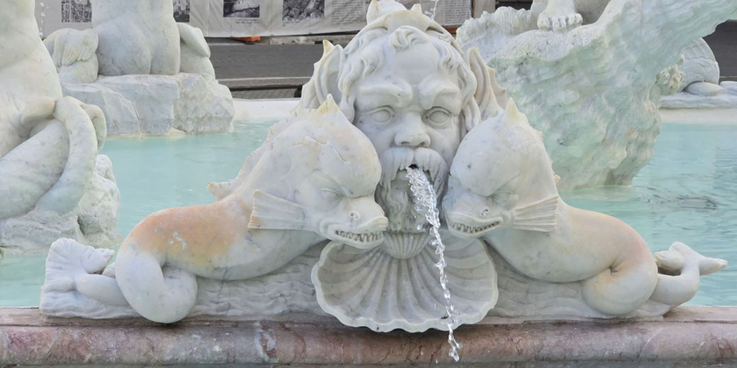 Ornate marble fountain featuring a bearded face and two sea creatures, all spouting water into a pool, with intricate shell and aquatic details.