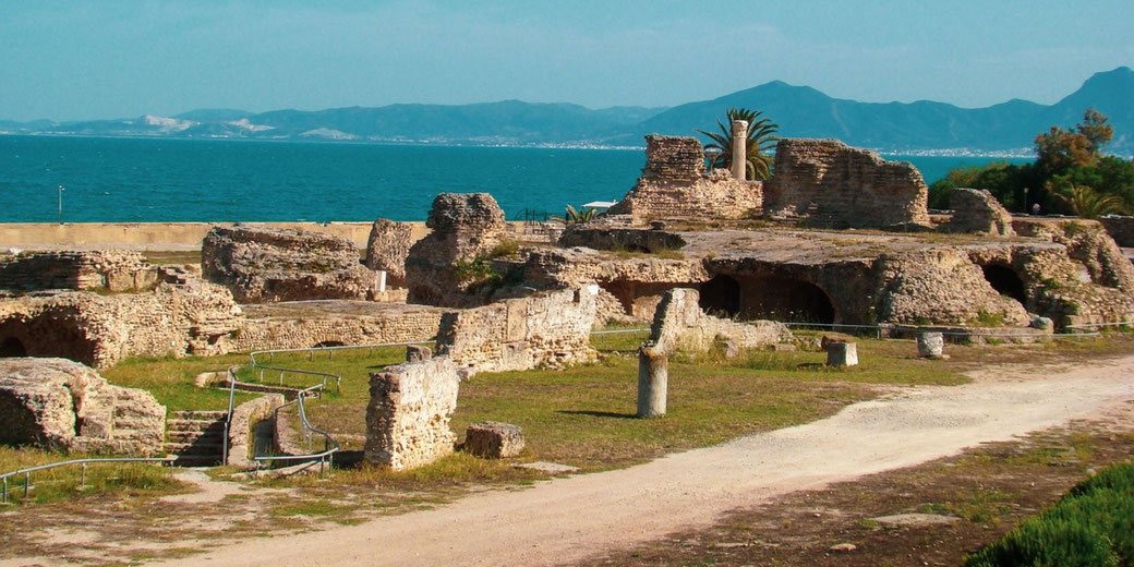 Ancient stone ruins overlooking a deep blue sea, with scattered columns and crumbling walls set against a backdrop of distant mountains.