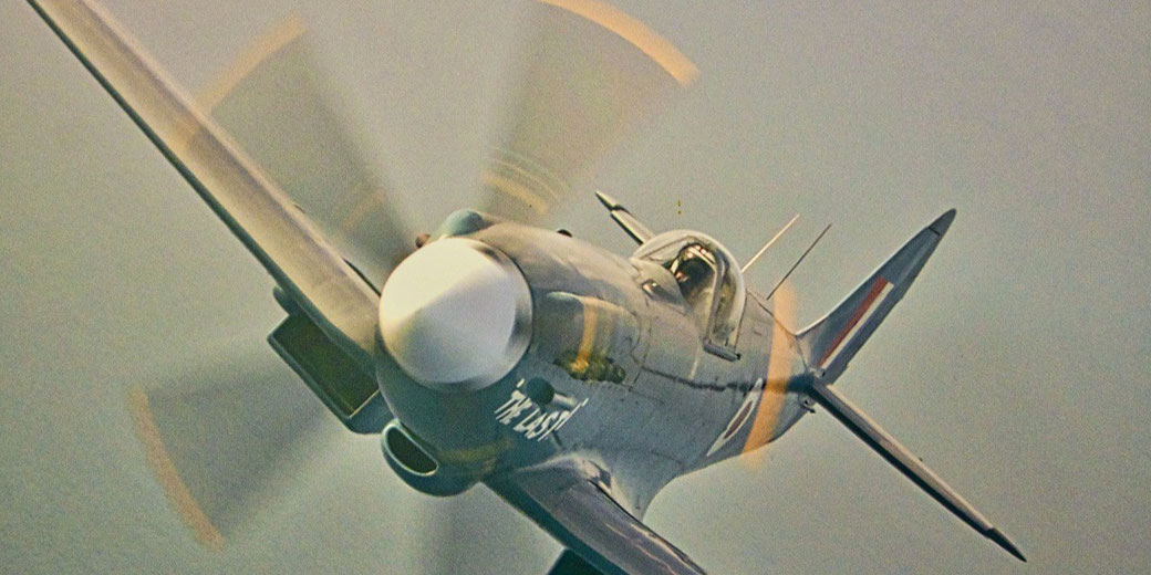 A vintage fighter plane with spinning propeller soars at an angle, captured mid-flight from a dramatic front view.