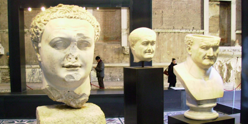 Three Roman marble busts of male figures are displayed on pedestals in a museum with ancient stone walls in the background.