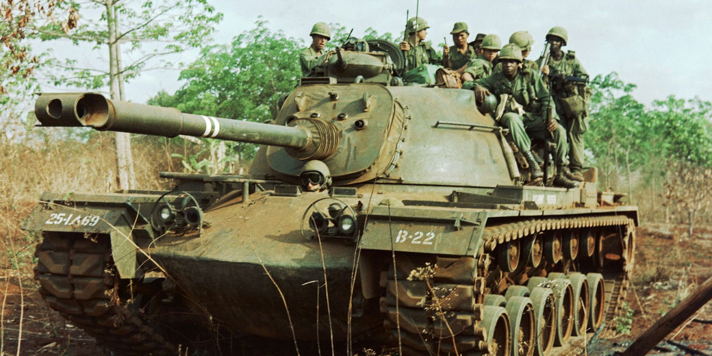 A group of soldiers ride atop a heavily armored tank in a rural, forested area during military operations.