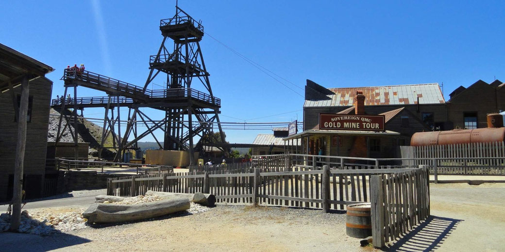 Old gold mine site with wooden buildings, headframe tower, fenced yard, and clear sky under bright sunlight in a dusty setting.